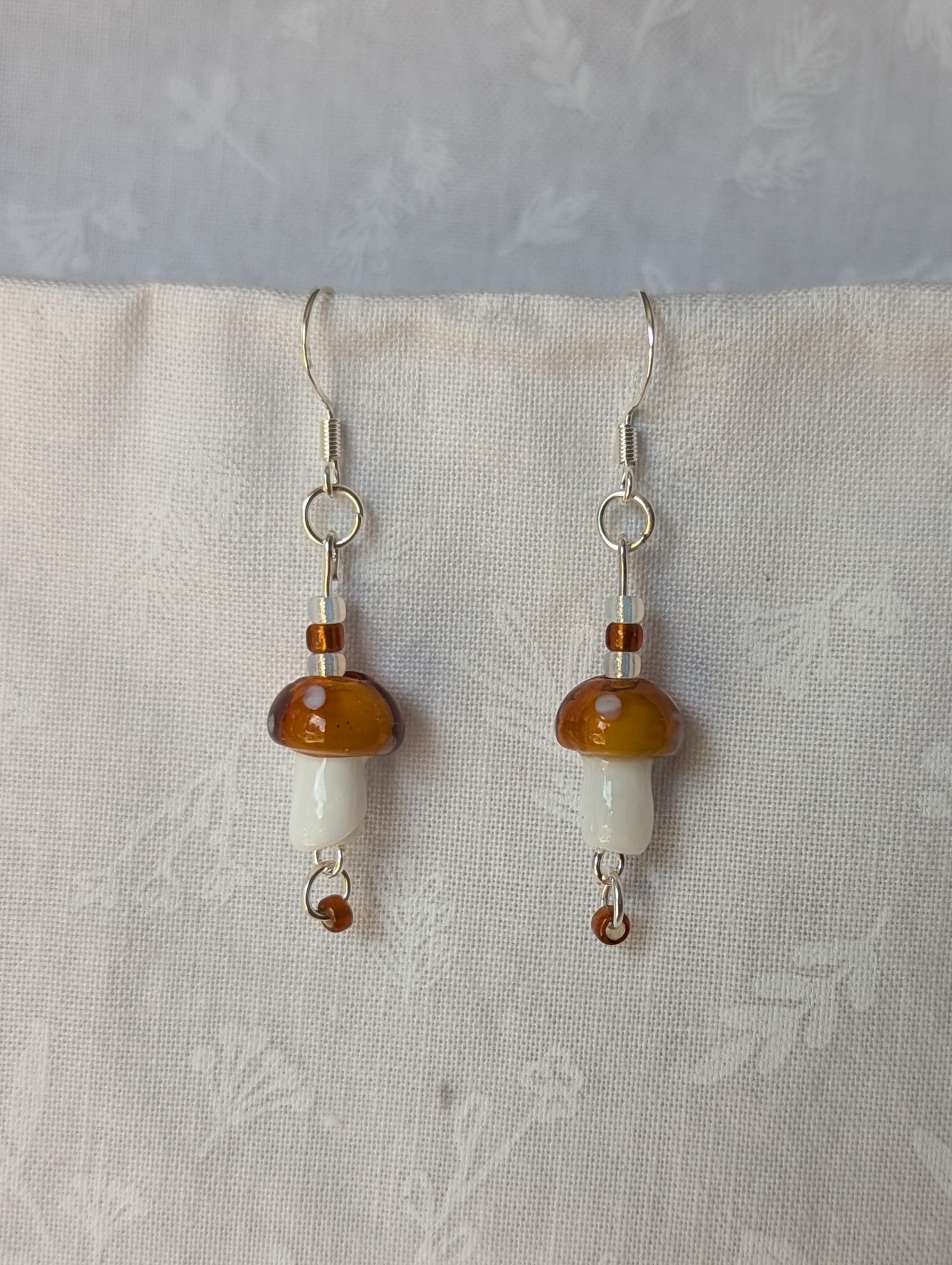 Handmade brown mushroom earrings with glass toadstools, brown and white seed beads, and sterling silver hooks.