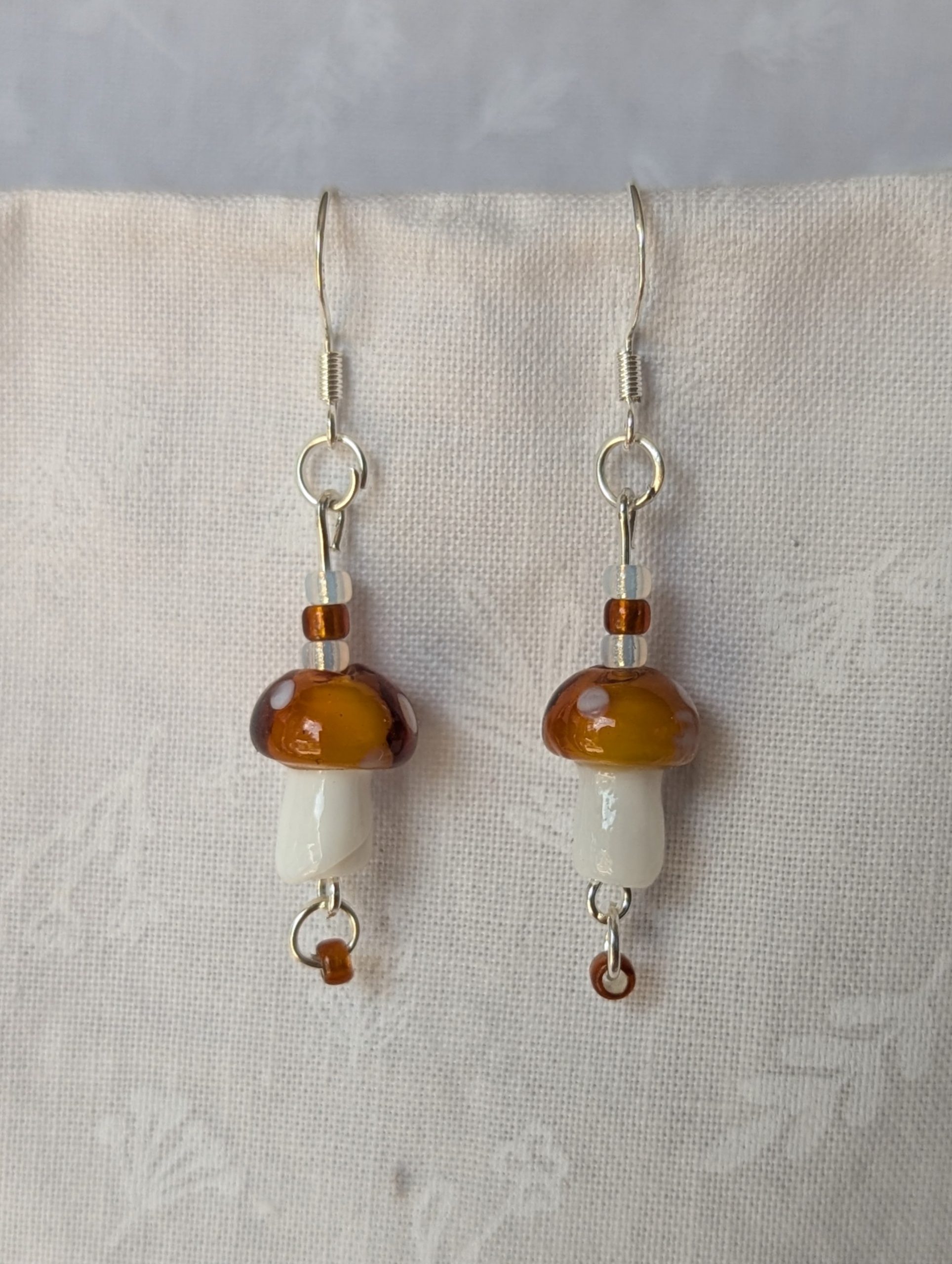 Handmade brown mushroom earrings with glass toadstools, brown and white seed beads, and sterling silver hooks.