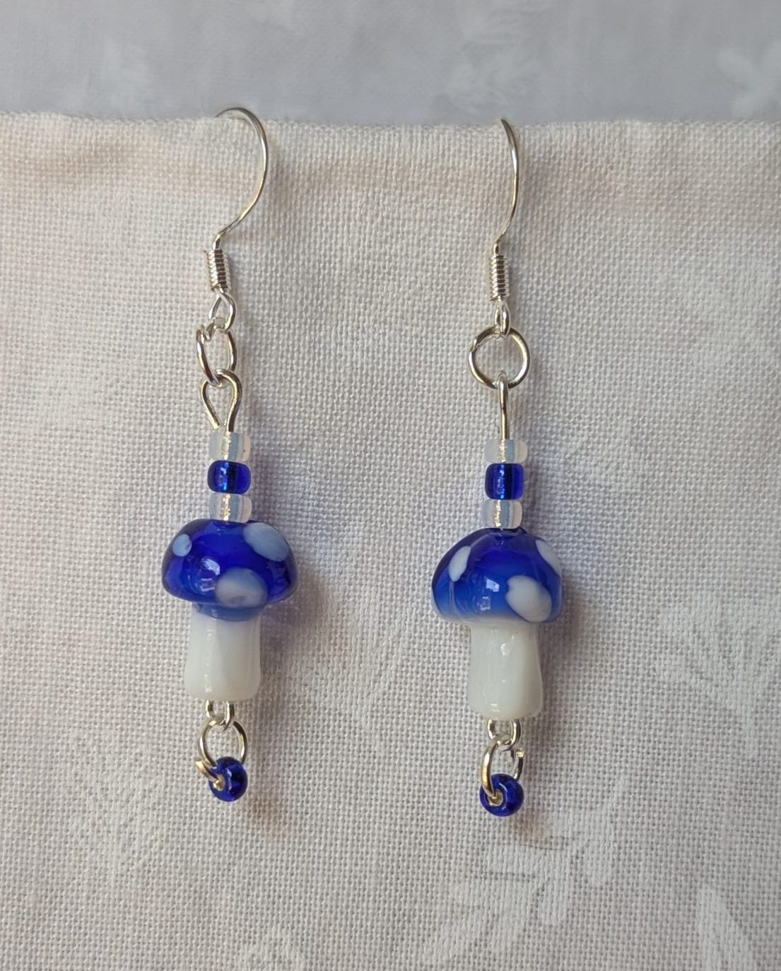 Handmade blue mushroom earrings with glass toadstools, blue and white seed beads, and sterling silver hooks.