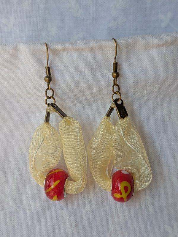 Handmade pale yellow ribbon earrings featuring bright red glass beads with yellow accents and hung from bronze-colored hooks.
