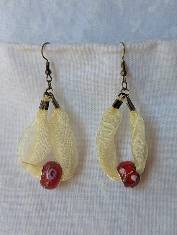 Handmade pale yellow ribbon earrings featuring red glass beads with shimmering gold accents and hung from bronze-colored hooks.