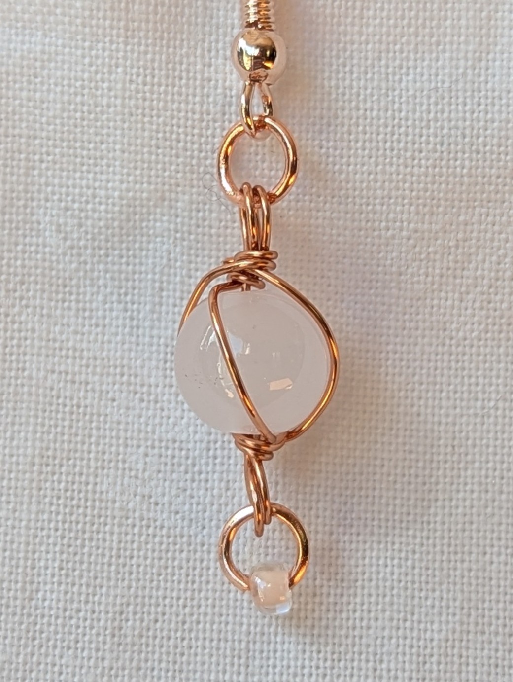 Handmade earrings featuring a milky white glass bead wrapped in copper wire with a white seed bead dangling below, hung from rose-gold colored hooks — Moonlit Copper Glow Earrings.