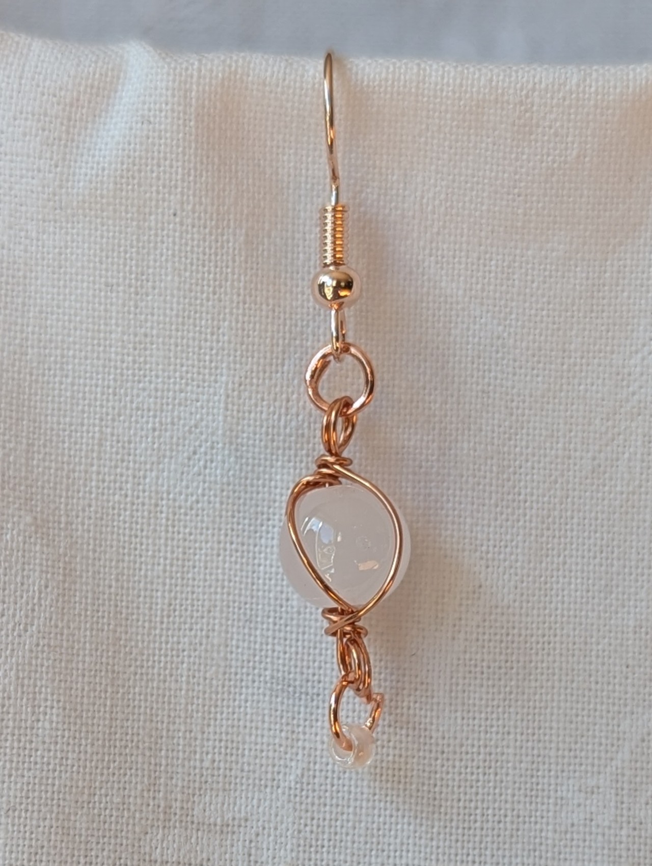 Handmade earrings featuring a milky white glass bead wrapped in copper wire with a white seed bead dangling below, hung from rose-gold colored hooks — Moonlit Copper Glow Earrings.
