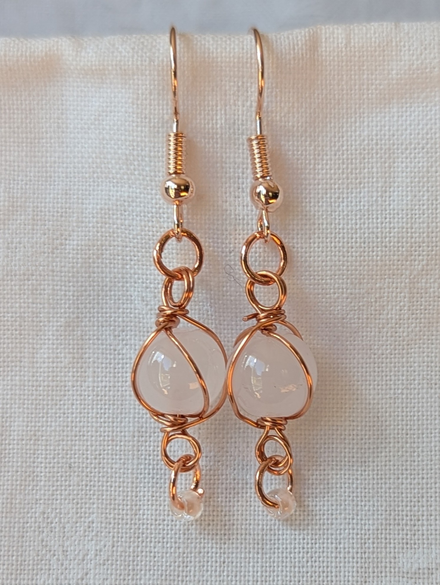 Handmade earrings featuring a milky white glass bead wrapped in copper wire with a white seed bead dangling below, hung from rose-gold colored hooks — Moonlit Copper Glow Earrings.