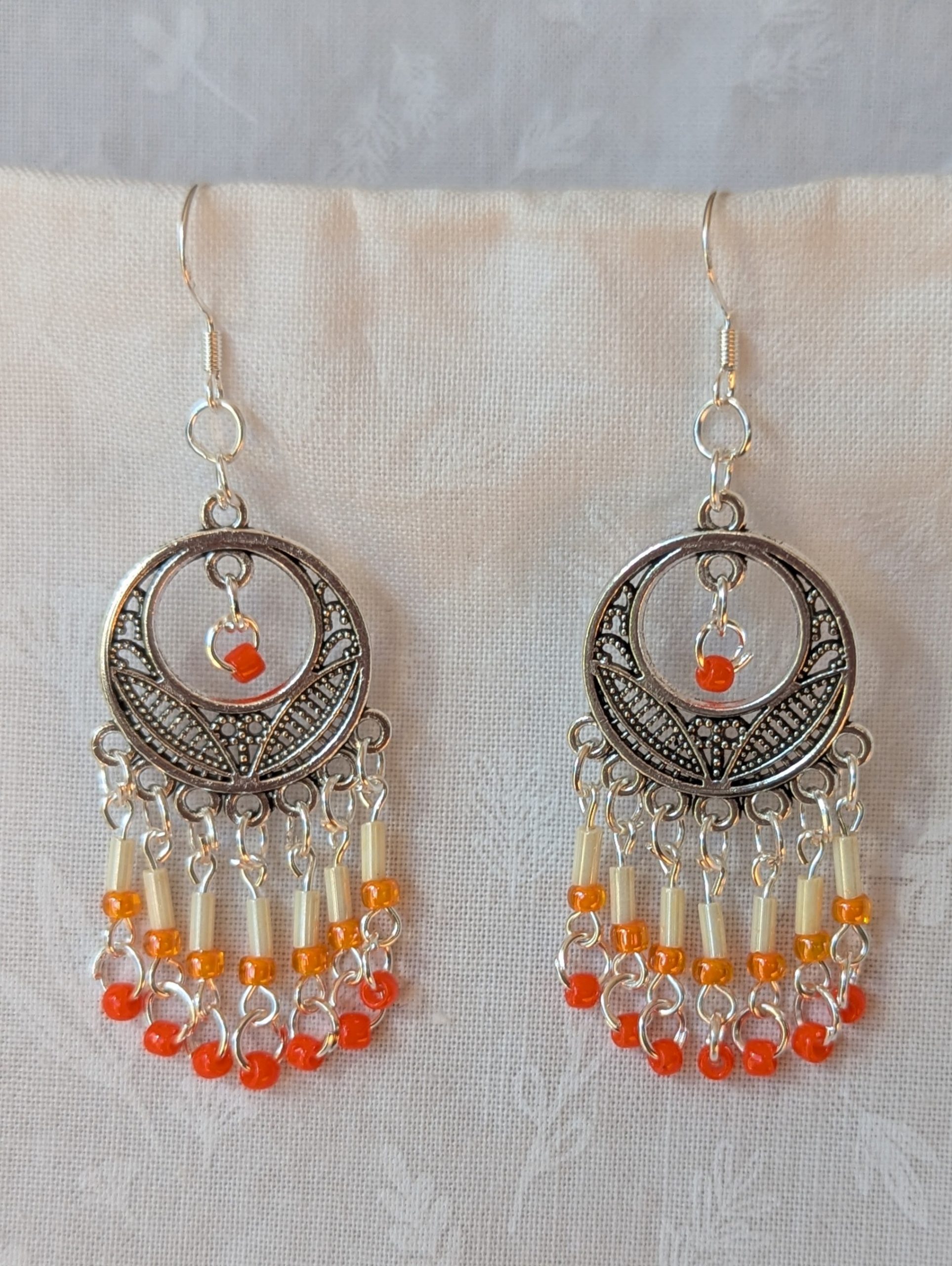 Orange and silver beaded decorative dangle earrings with cream, pale orange, and dark orange glass beads hanging in cascading strands from a silver ornamental medallion.