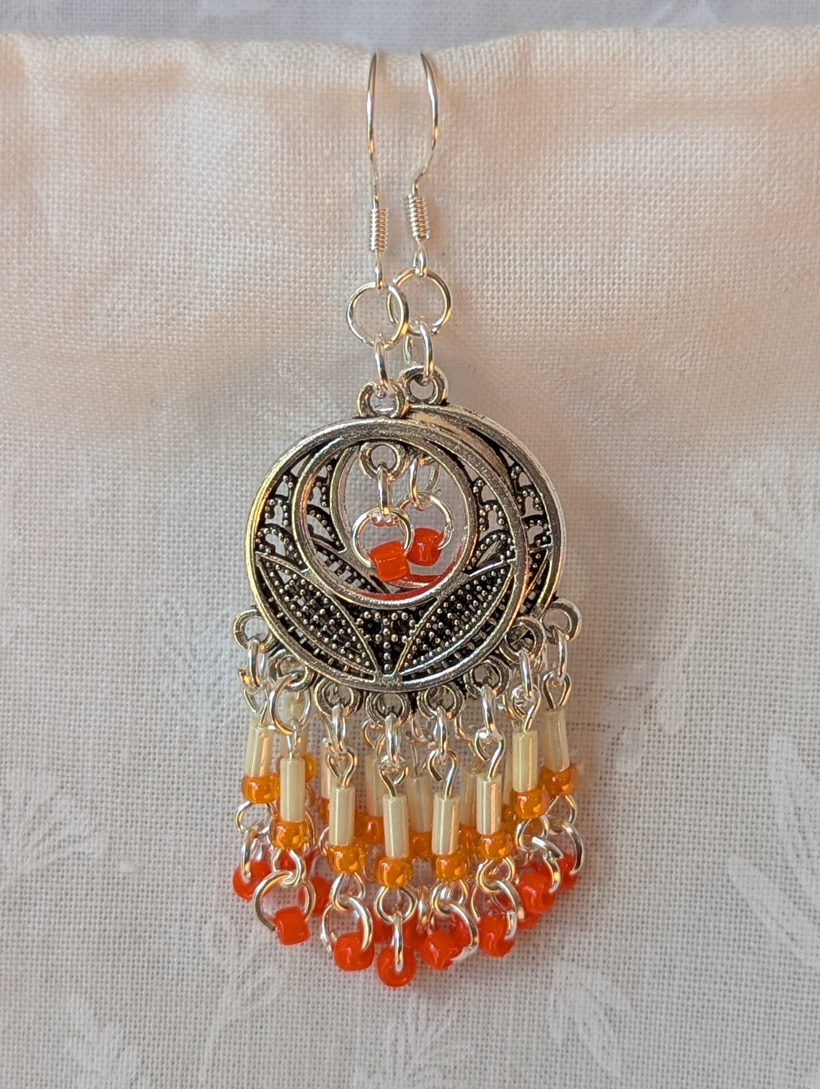 Orange and silver beaded decorative dangle earrings with cream, pale orange, and dark orange glass beads hanging in cascading strands from a silver ornamental medallion.