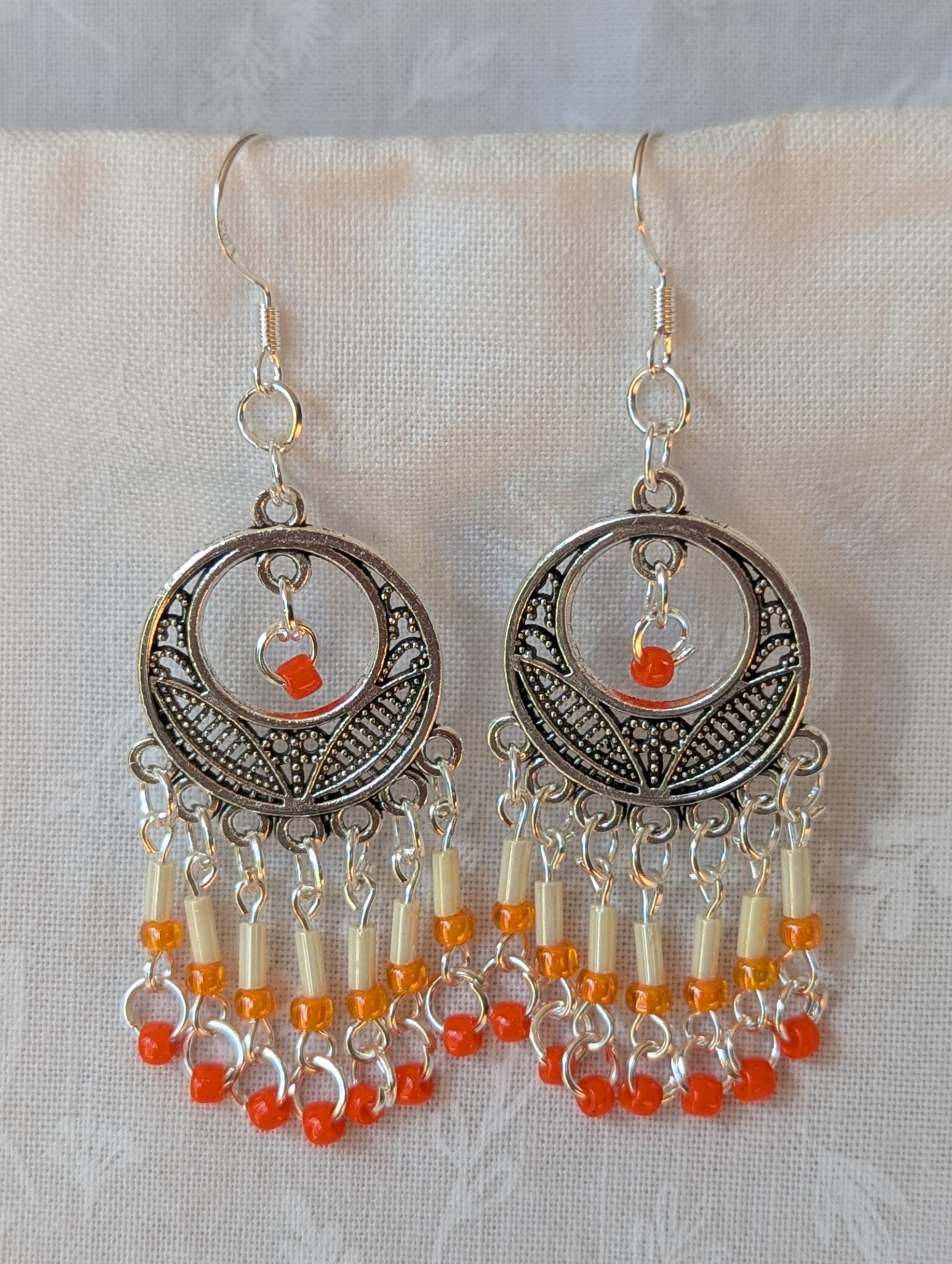 Orange and silver beaded decorative dangle earrings with cream, pale orange, and dark orange glass beads hanging in cascading strands from a silver ornamental medallion.