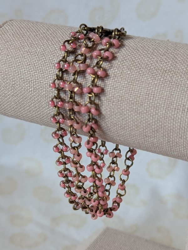 Triple-strand bronze chain bracelet with pale pink beads in pearly, iridescent, and lined clear finishes, displayed on a fabric bracelet stand.