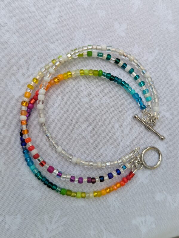 Triple-strand handmade bracelet with rainbow seed beads, pearl-white and clear glass beads, steel-colored memory wire, and toggle clasp closure.