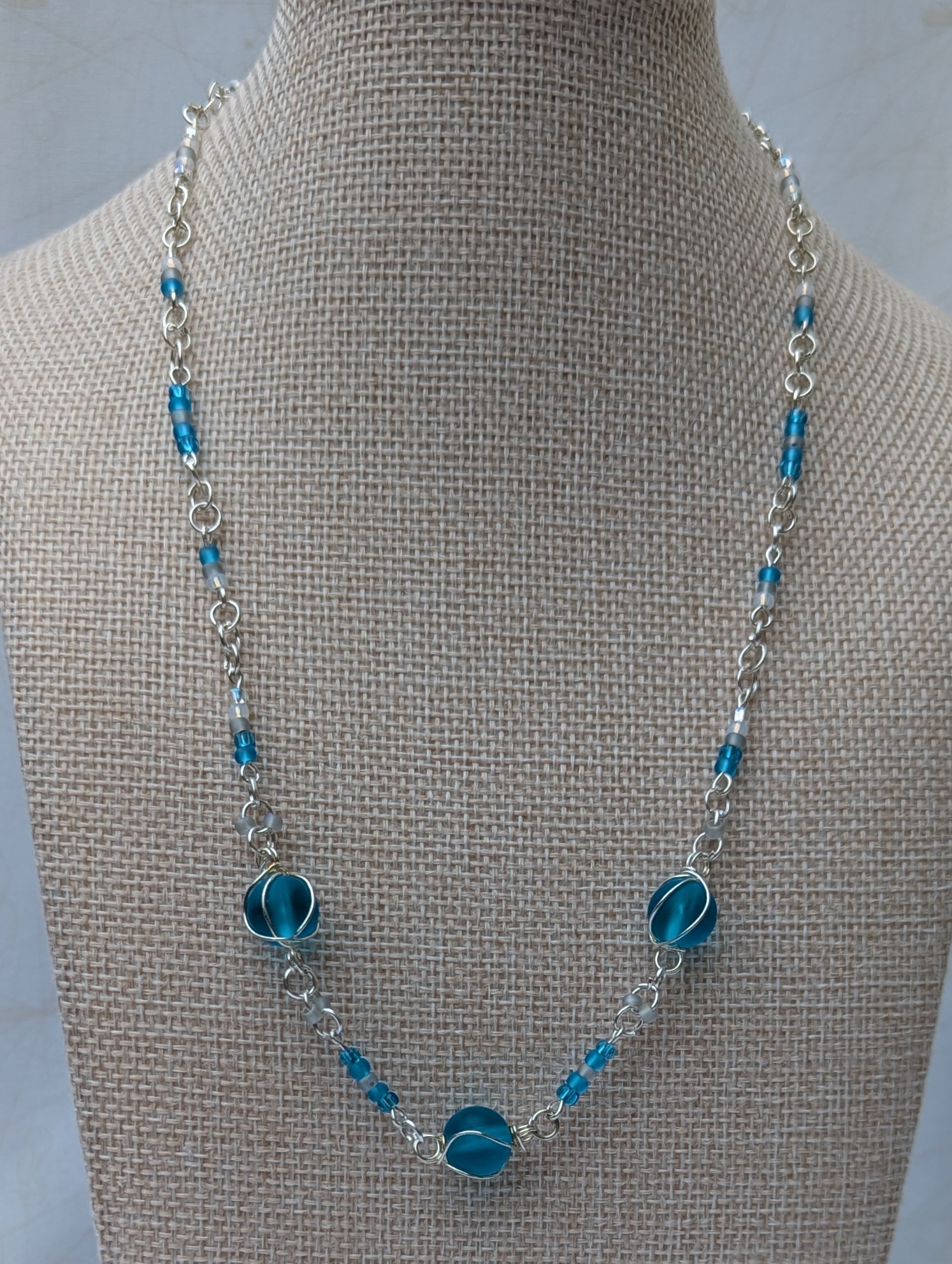 Handmade silver-colored chain necklace with pale aqua and frosted white seed beads, featuring three wire-wrapped ocean-blue glass beads and a silver lobster clasp closure.