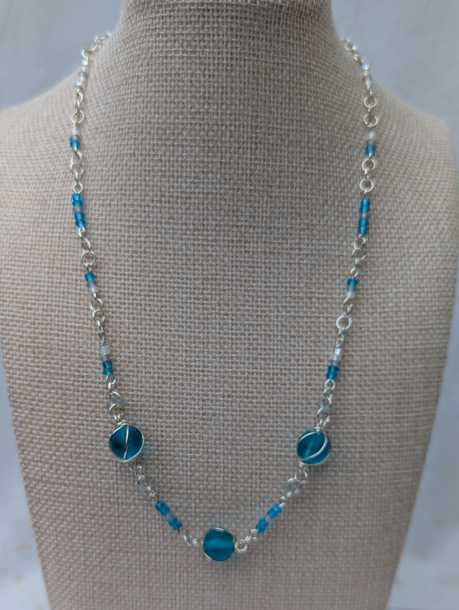 Handmade silver-colored chain necklace with pale aqua and frosted white seed beads, featuring three wire-wrapped ocean-blue glass beads and a silver lobster clasp closure.