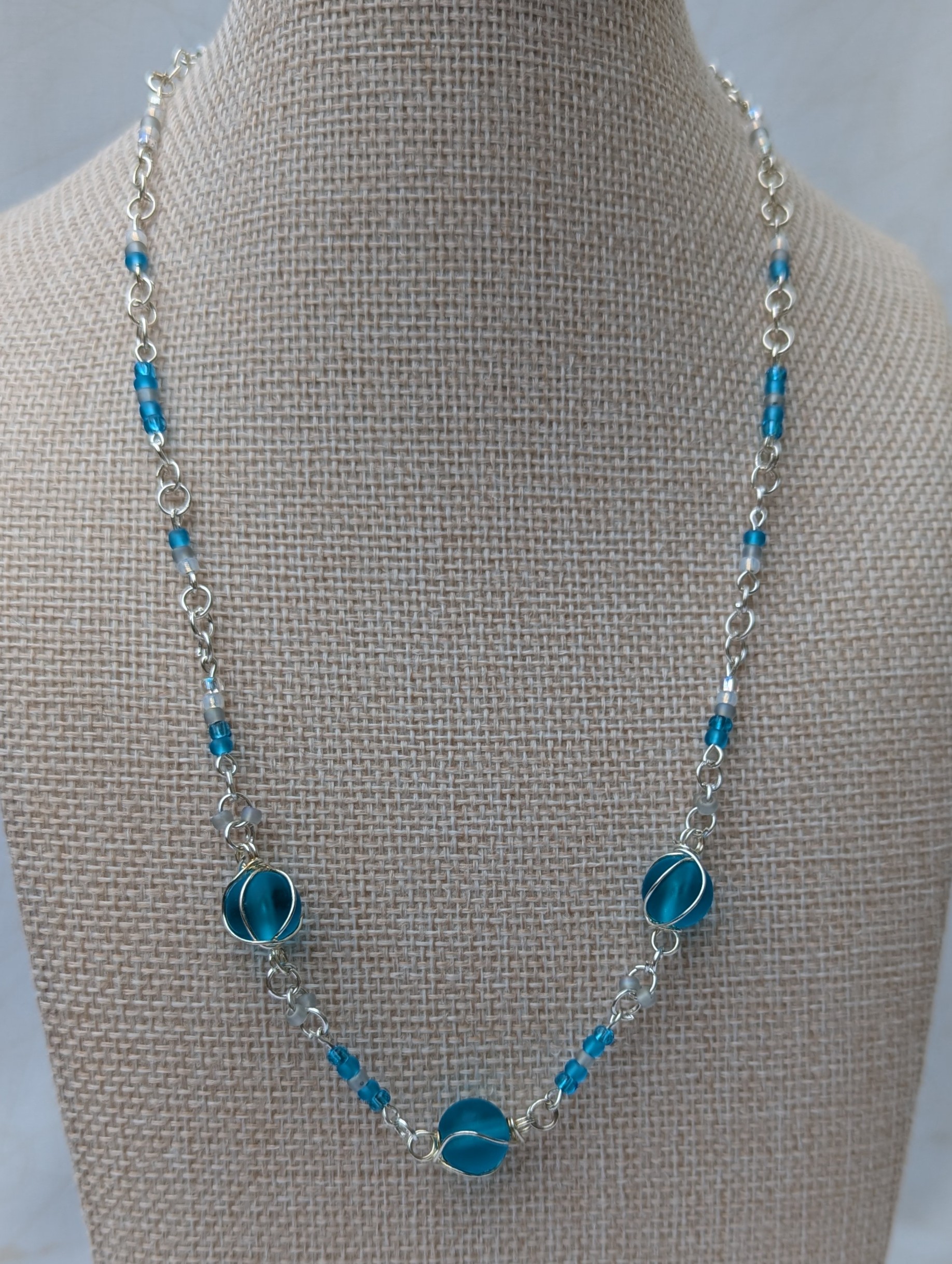 Handmade silver-colored chain necklace with pale aqua and frosted white seed beads, featuring three wire-wrapped ocean-blue glass beads and a silver lobster clasp closure.