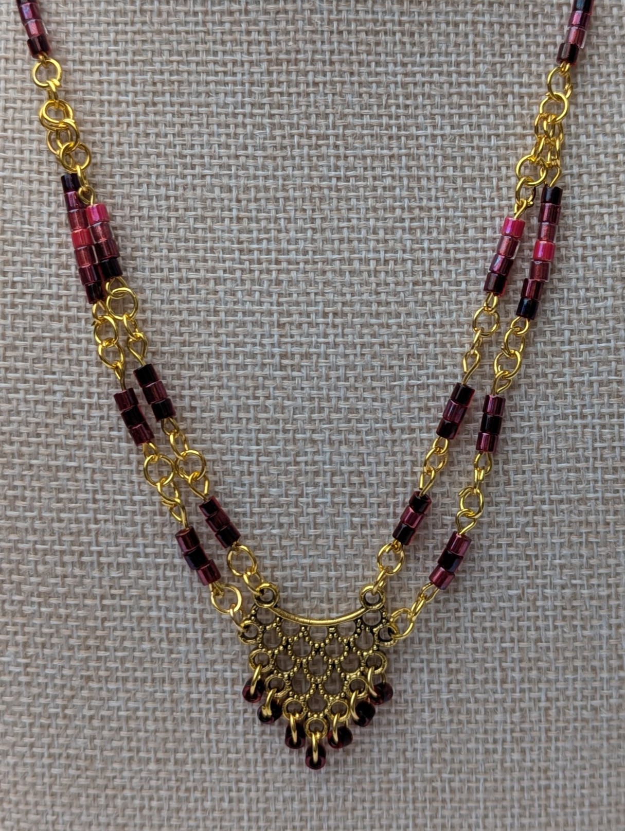 Handmade gold-toned double chain necklace with garnet, burgundy, and deep wine glass seed beads, featuring an ornate gold pendant and lobster clasp closure.