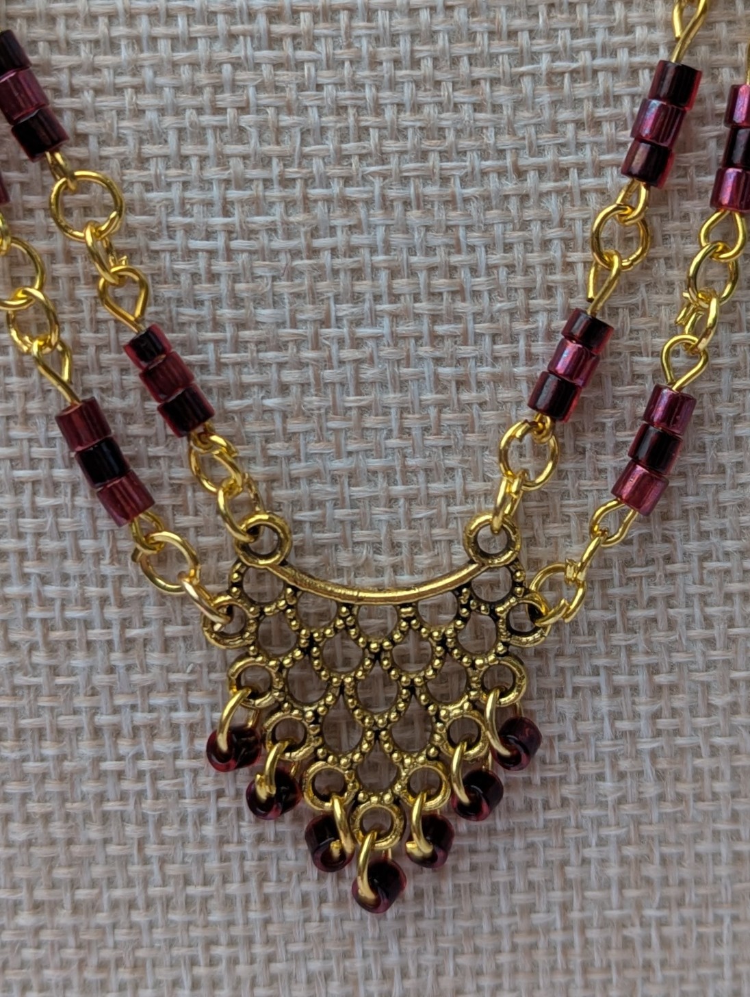 Handmade gold-toned double chain necklace with garnet, burgundy, and deep wine glass seed beads, featuring an ornate gold pendant and lobster clasp closure.