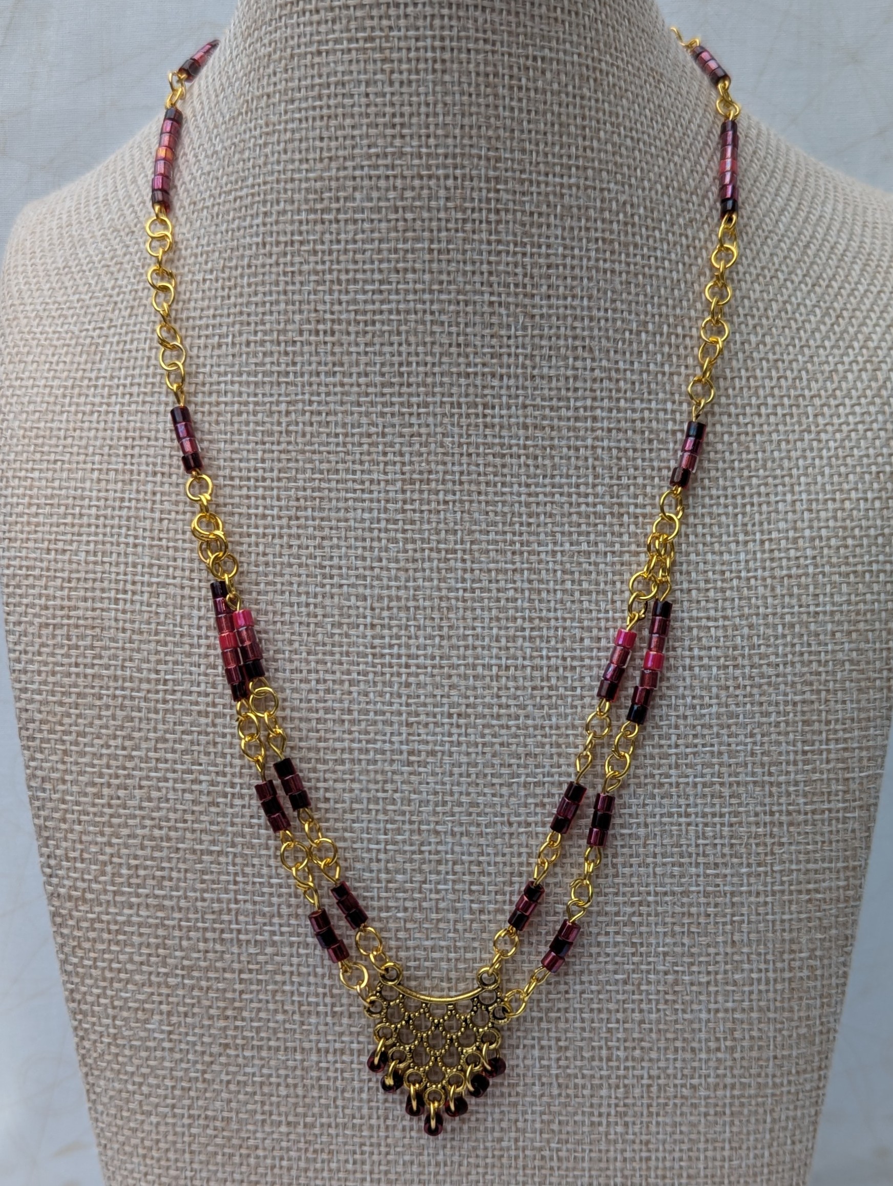 Handmade gold-toned double chain necklace with garnet, burgundy, and deep wine glass seed beads, featuring an ornate gold pendant and lobster clasp closure.